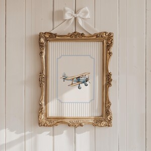 May include: A framed print of a blue and white airplane with brown wooden wings. The frame is gold with ornate detailing and is hanging on a white wood wall.