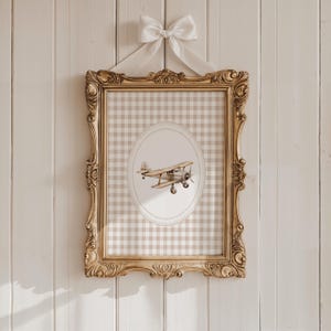 Pu&ograve; includere: Un'opera d'arte incorniciata con un'illustrazione di aereo vintage. La cornice dorata e ornata circonda uno sfondo a quadretti gingham. Un fiocco bianco &egrave; legato in alto. L'opera &egrave; pensata per l'arredamento della cameretta.