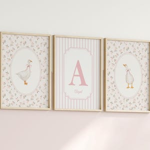 May include: Three framed wall art prints with light wood frames. The two outer prints feature a white goose with a pink bow inside an oval frame against a floral background. The center print displays a large pink letter A and the name Abigail.