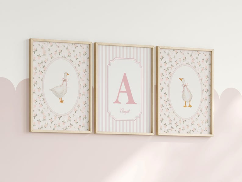 May include: Three framed wall art prints with light wood frames. The two outer prints feature a white goose with a pink bow inside an oval frame against a floral background. The center print displays a large pink letter A and the name Abigail.
