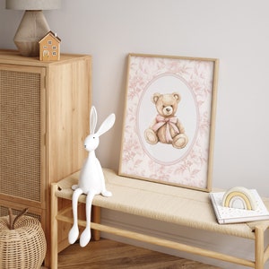 Pu&ograve; includere: Una stampa ad acquerello di un orsacchiotto con un fiocco rosa, incorniciata in una cornice di legno chiaro, con uno sfondo a motivi floreali. La stampa &egrave; appesa a un muro bianco sopra una panca di legno chiaro con una statuetta di coniglio bianco seduta sopra.