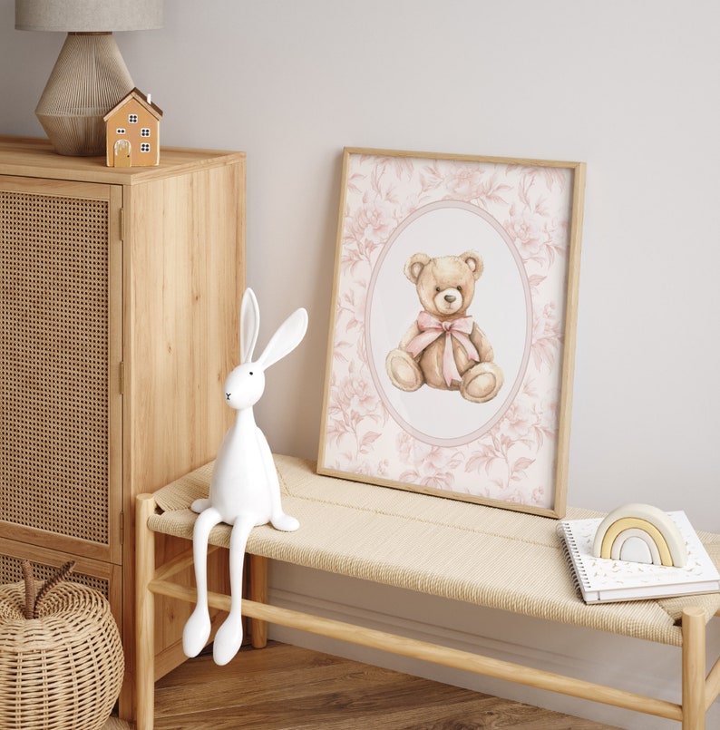 Pu&ograve; includere: Una stampa ad acquerello di un orsacchiotto con un fiocco rosa, incorniciata in una cornice di legno chiaro, con uno sfondo a motivi floreali. La stampa &egrave; appesa a un muro bianco sopra una panca di legno chiaro con una statuetta di coniglio bianco seduta sopra.