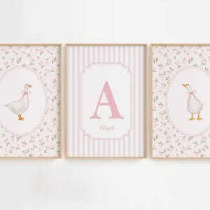 May include: A set of three framed wall art prints. The two outer prints feature a white goose with a pink ribbon in an oval frame against a floral background. The center print displays a large pink letter A and the name Abigail in a striped frame.