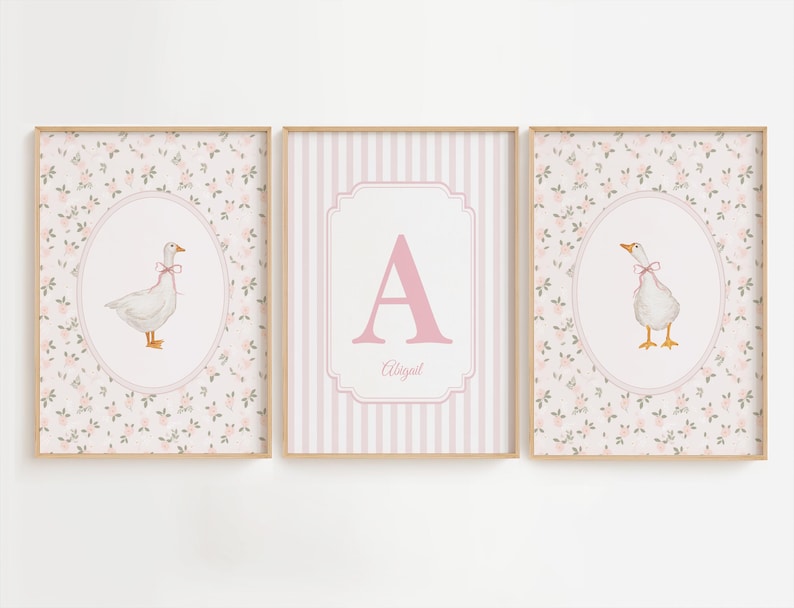 May include: A set of three framed wall art prints. The two outer prints feature a white goose with a pink ribbon in an oval frame against a floral background. The center print displays a large pink letter A and the name Abigail in a striped frame.