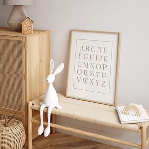 May include: A white rabbit figurine with long ears sits on a light wood bench. A framed alphabet print and a wooden rainbow decor are also on the bench. A wooden cabinet and a small house figurine are in the background.
