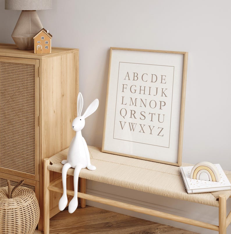 May include: A white rabbit figurine with long ears sits on a light wood bench. A framed alphabet print and a wooden rainbow decor are also on the bench. A wooden cabinet and a small house figurine are in the background.