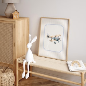 May include: A white rabbit figurine with long legs sits on a wooden bench in front of a framed print of a blue airplane against a striped background.