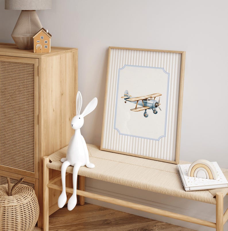 May include: A white rabbit figurine with long legs sits on a wooden bench in front of a framed print of a blue airplane against a striped background.
