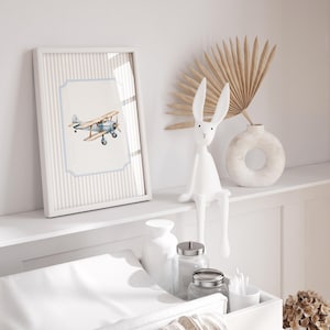 May include: A white changing table with a white changing pad, a white ceramic vase with a dried palm leaf, a white ceramic ring-shaped vase, a white ceramic bunny figurine, and a framed print of a blue and white airplane on a striped background.