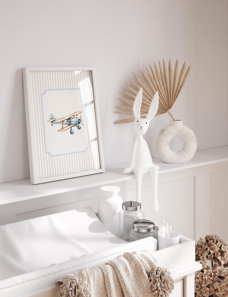 May include: A white changing table with a white changing pad, a white ceramic vase with a dried palm leaf, a white ceramic ring-shaped vase, a white ceramic bunny figurine, and a framed print of a blue and white airplane on a striped background.