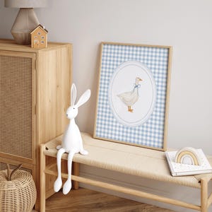 May include: A white goose with a blue bow sits in a circle on a blue and white gingham background. The goose is surrounded by a white lace border. The image is framed in a light wood frame.