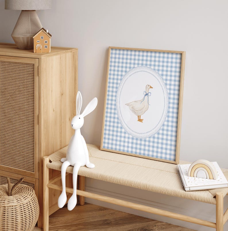 May include: A white goose with a blue bow sits in a circle on a blue and white gingham background. The goose is surrounded by a white lace border. The image is framed in a light wood frame.