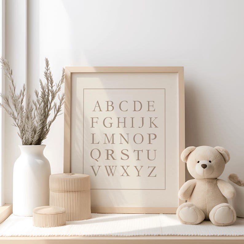 May include: A framed alphabet print with a beige background and light brown letters. A beige teddy bear sits to the right of the frame. A white vase with dried plants and two beige decorative boxes are on the left.