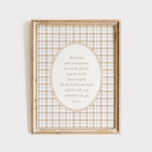 May include: Framed wall art with a beige and white gingham pattern background. An oval shape in the center contains text from Joshua 1:9, in a soft, elegant font. The frame is a light brown color.
