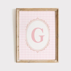 May include: Framed wall art featuring a pink gingham pattern background. A light pink letter "G" is centered within an oval frame with ornate detailing. The frame is a light wood color.