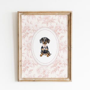 May include: Framed art print featuring a watercolor illustration of a dachshund dog with a pink bow tie. The dog is centered within an oval frame, set against a floral patterned background in shades of pink and cream. The frame is a light, warm wood tone.