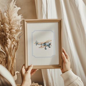 May include: A framed print of a blue and brown vintage airplane with a light blue background and white stripes. The frame is made of wood and has a light brown finish.