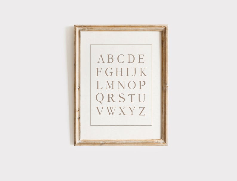 May include: A framed alphabet print. The print features the letters of the alphabet in a serif font, arranged in rows. The letters are a muted brown color against a cream background. The frame is a light brown, with a slightly distressed look.