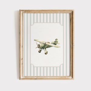May include: A framed print featuring a vintage-style airplane illustration. The airplane is painted in shades of green and tan, set against a white background with a decorative border. The frame has a light wood finish and a striped background.