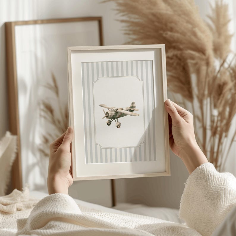 May include: A framed print featuring a vintage-style airplane illustration. The artwork is set against a backdrop of vertical gray and white stripes, within a white frame. The airplane is painted in shades of green and brown.