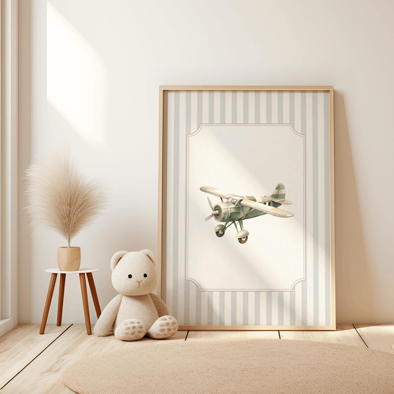 May include: A framed print featuring a watercolor illustration of a vintage airplane in shades of green and cream. The artwork is set against a backdrop of vertical gray and white stripes, framed in light wood. A teddy bear and a small table with a plant are in the foreground.