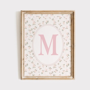 Può includere: Stampa floreale incorniciata con una grande lettera "M" rosa all'interno di un ovale. Lo sfondo presenta piccoli fiori rosa e foglie verdi su sfondo bianco. La cornice è color legno chiaro.