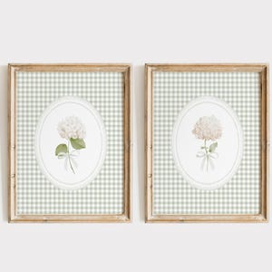 May include: Two framed floral prints. Each print features a white hydrangea flower with green leaves and a ribbon, set against a light green and white gingham background. The frames are a light brown color.