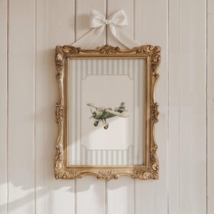 May include: A framed print featuring a vintage-style airplane. The print is set within a gold ornate frame and is suspended by a white bow. The background of the print has a striped pattern in light blue and white.
