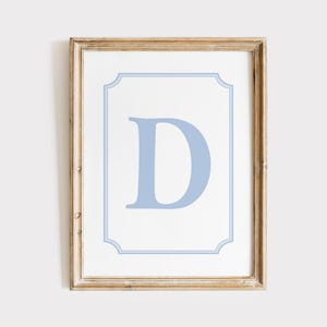 Puede incluir: Arte de pared enmarcado con una gran letra "D" azul claro sobre un fondo blanco. La letra está centrada dentro de un borde decorativo azul claro. El marco es de color marrón claro.