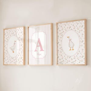 May include: Three framed wall art prints with light wood frames. The outer two prints feature a white goose with a pink bow on a floral background inside an oval border. The center print displays a large pink letter A and the name Abigail.