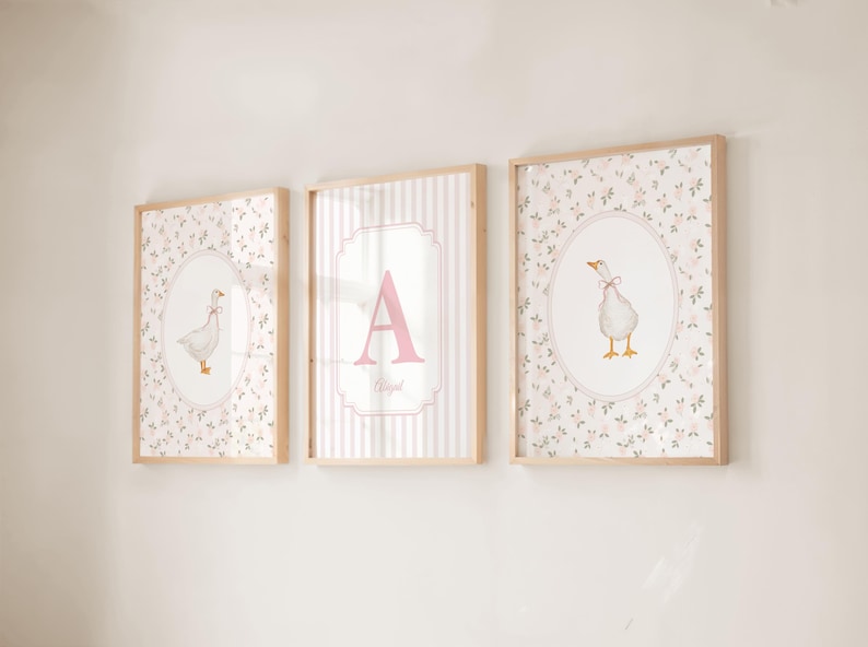 May include: Three framed wall art prints with light wood frames. The outer two prints feature a white goose with a pink bow on a floral background inside an oval border. The center print displays a large pink letter A and the name Abigail.