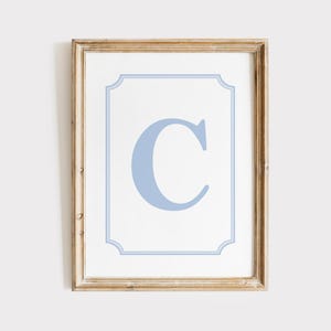 May include: Framed print featuring a light blue letter "C" on a white background, framed by a light blue border. The frame is a light brown color.