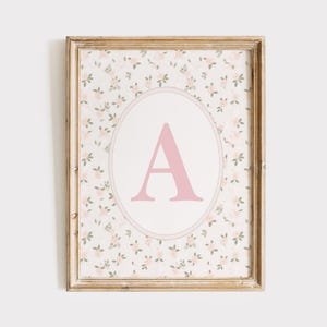 Op de afbeelding: Een ingelijste print met een bloemenpatroon achtergrond en een ovale midden. Het midden toont een grote, roze letter "A". De lijst is licht, verweerd hout. Het bloemenpatroon bevat kleine roze bloemen en groene bladeren.