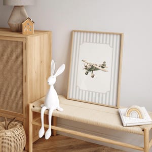 May include: A framed print of a vintage airplane with a light gray and white striped background. The print is in a light wood frame. A white rabbit figurine sits on a woven bench, next to a wooden cabinet and a small wooden house.
