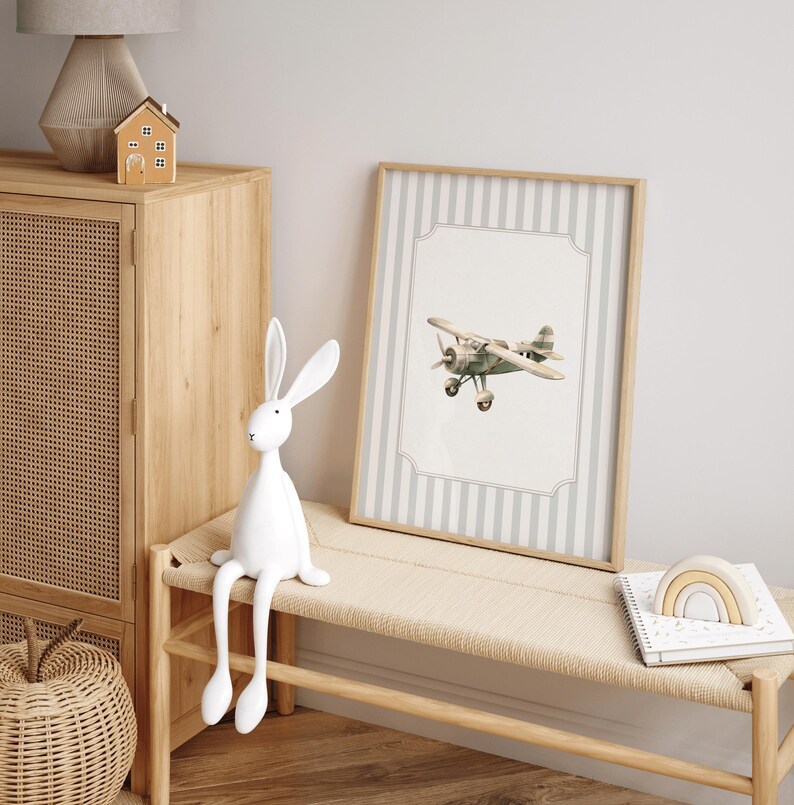 May include: A framed print of a vintage airplane with a light gray and white striped background. The print is in a light wood frame. A white rabbit figurine sits on a woven bench, next to a wooden cabinet and a small wooden house.