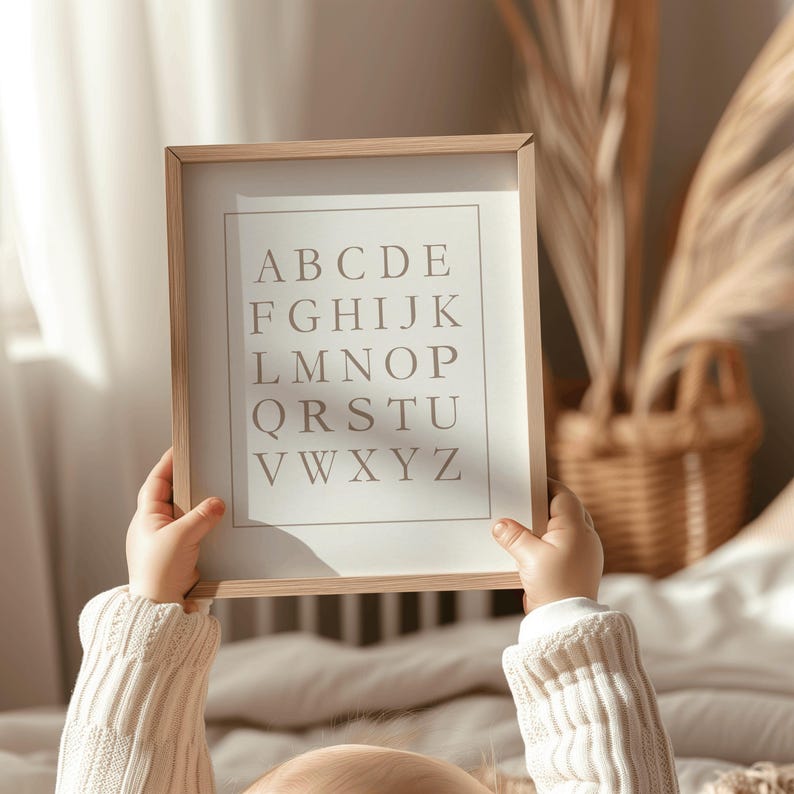 May include: A framed print featuring the alphabet in a neutral color palette. The letters are arranged in rows, with each letter clearly visible. The frame is a light wood tone, and the print is held by a person wearing a cream-colored sweater.