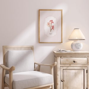 May include: A white lamp with a pleated white shade sits on a light wood side table with a drawer and a cabinet door. A framed print with pink flowers and a striped background hangs on the wall behind the table. A light wood chair with a white cushion sits in the foreground.
