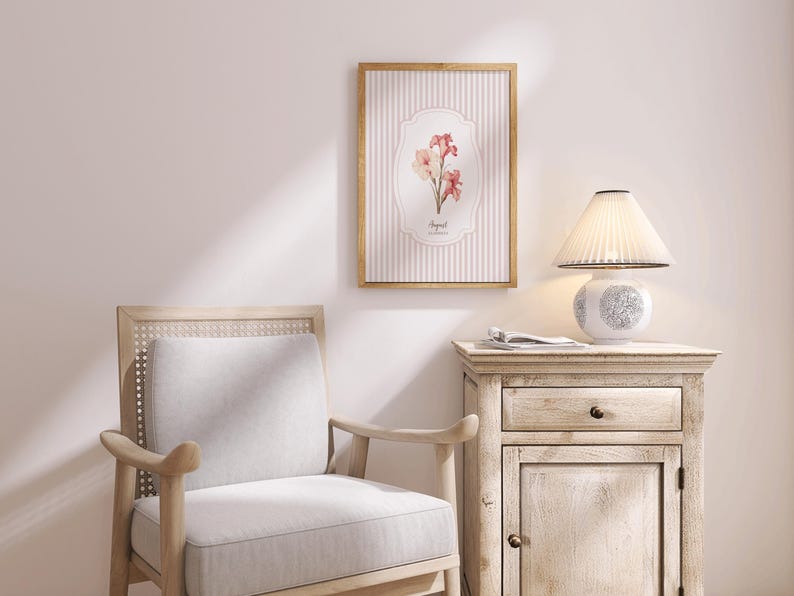 May include: A white lamp with a pleated white shade sits on a light wood side table with a drawer and a cabinet door. A framed print with pink flowers and a striped background hangs on the wall behind the table. A light wood chair with a white cushion sits in the foreground.