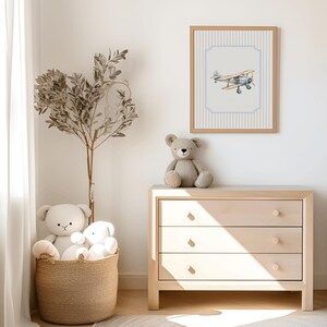 May include: A light wood dresser with three drawers, a teddy bear sitting on top, and a framed print of a blue airplane on a white and blue striped background.