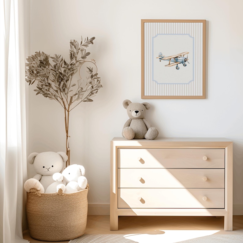 May include: A light wood dresser with three drawers, a teddy bear sitting on top, and a framed print of a blue airplane on a white and blue striped background.