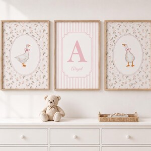 May include: Three framed wall art prints featuring a light pink and white color palette. The two outer prints display a white goose with a pink bow inside an oval, while the center print features a large pink letter A and the name Abigail.