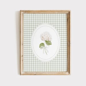 Op de afbeelding: Een ingelijste print met een aquarel illustratie van een witte hortensia bloem, groene bladeren en een wit lint. Het kunstwerk staat tegen een lichtgroen en wit ruitjespatroon, in een houten frame.