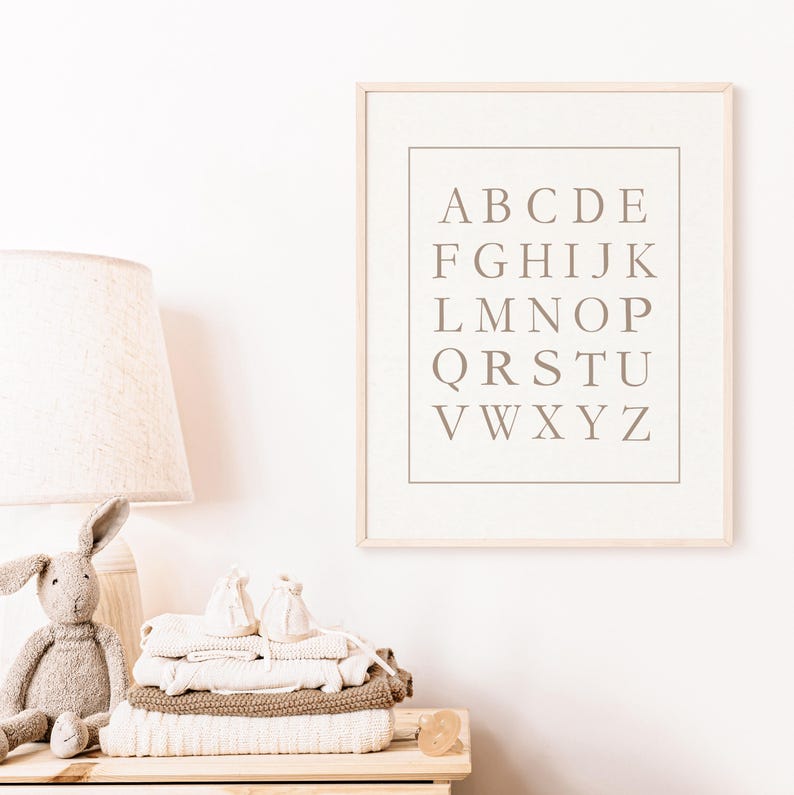 May include: A framed alphabet print in a light wood frame hangs on a white wall. The print features the letters A through Z in a serif font. A stack of folded baby clothes, a stuffed bunny, and a lamp sit on a wooden dresser.