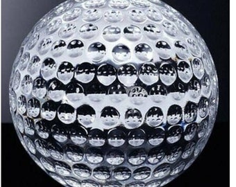Może przedstawiać: Przezroczysta szklana piłka golfowa z powierzchnią w kształcie dołków. Piłka leży na czarnej powierzchni i odbija światło.
