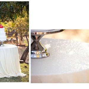 May include: A white three-tiered wedding cake with red flowers on top, sitting on a silver cake stand. The cake is on a white tablecloth with a lace trim. The cake stand is on a white table with a white tablecloth.