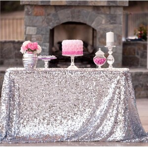 May include: A table with a silver sequined tablecloth, decorated with a pink ombre cake, flowers, and other decorative items. The table is set up outdoors with a stone fireplace in the background.