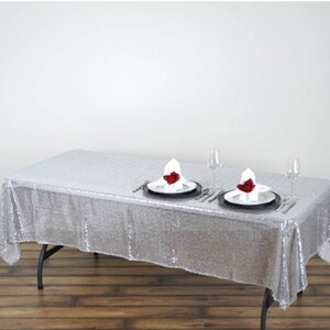 May include: A silver sequin tablecloth with a shimmering, sparkly finish. Two place settings are set on the table with white napkins and red roses.