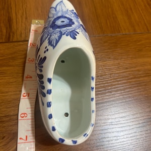 May include: A white ceramic Dutch clog planter with blue floral designs. The planter is approximately 8 inches long. The interior is light green. The clog has two holes on the side and is placed on a wooden surface.