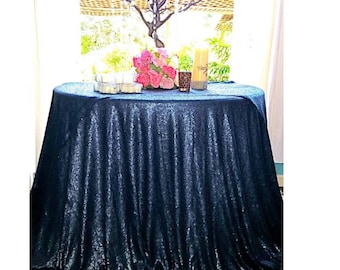 May include: A round table covered with a navy blue sequin tablecloth. The table is adorned with a floral arrangement, candles, and other decorative items. The table is positioned in front of a window.