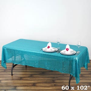 May include: A rectangular turquoise sequin tablecloth, measuring 60 x 102 inches, covers a black folding table. Two place settings with white plates, silverware, and folded napkins with a red rose are visible. Wine glasses complete the elegant setup.